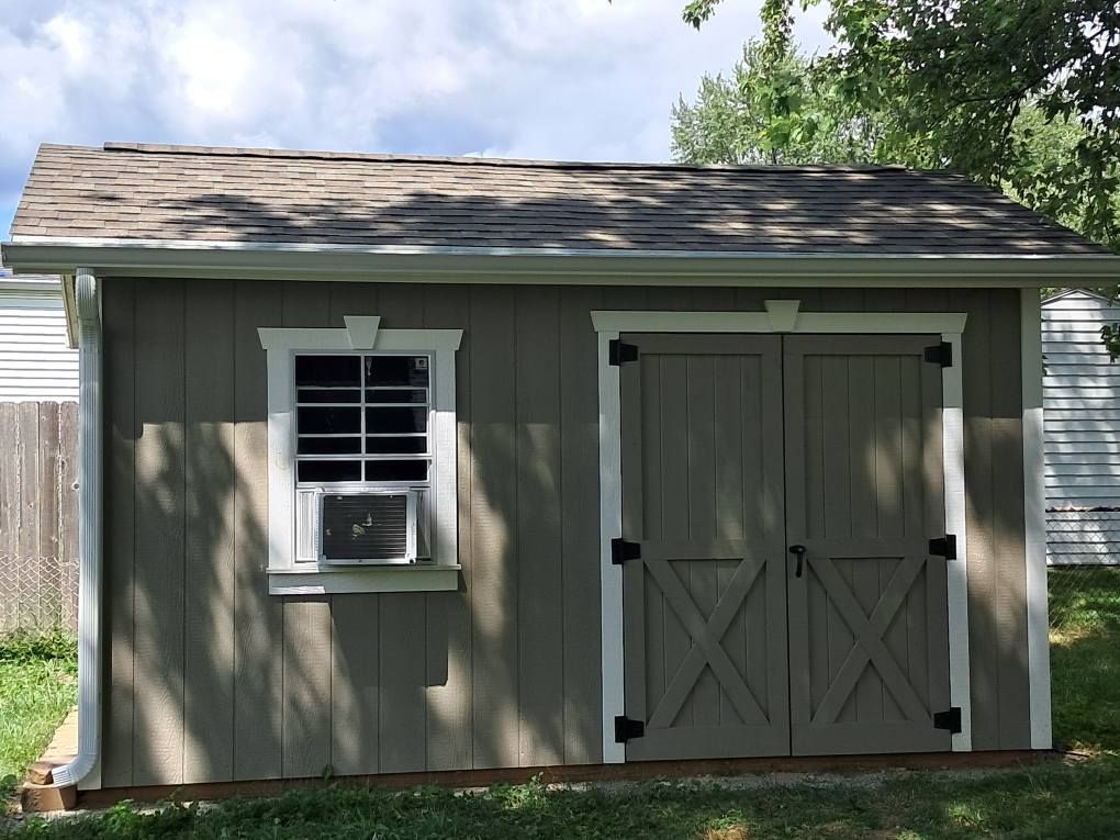 Tan shed with white trim, brown roof, and black door hardware. A window with an air conditioner is visible.
