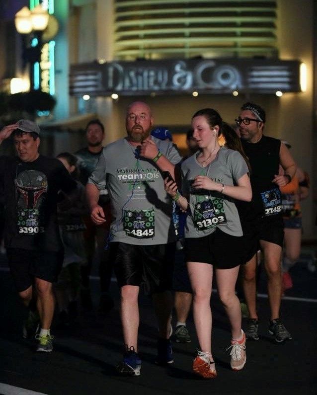 Runners at night race near DeSisto & Co sign. Man in grey shirt helps a woman; others run behind.