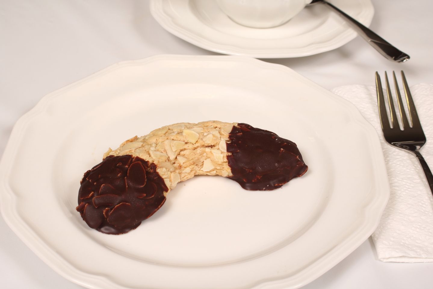 Almond crescent cookie dipped in chocolate on a white plate with fork, napkin, and teacup.