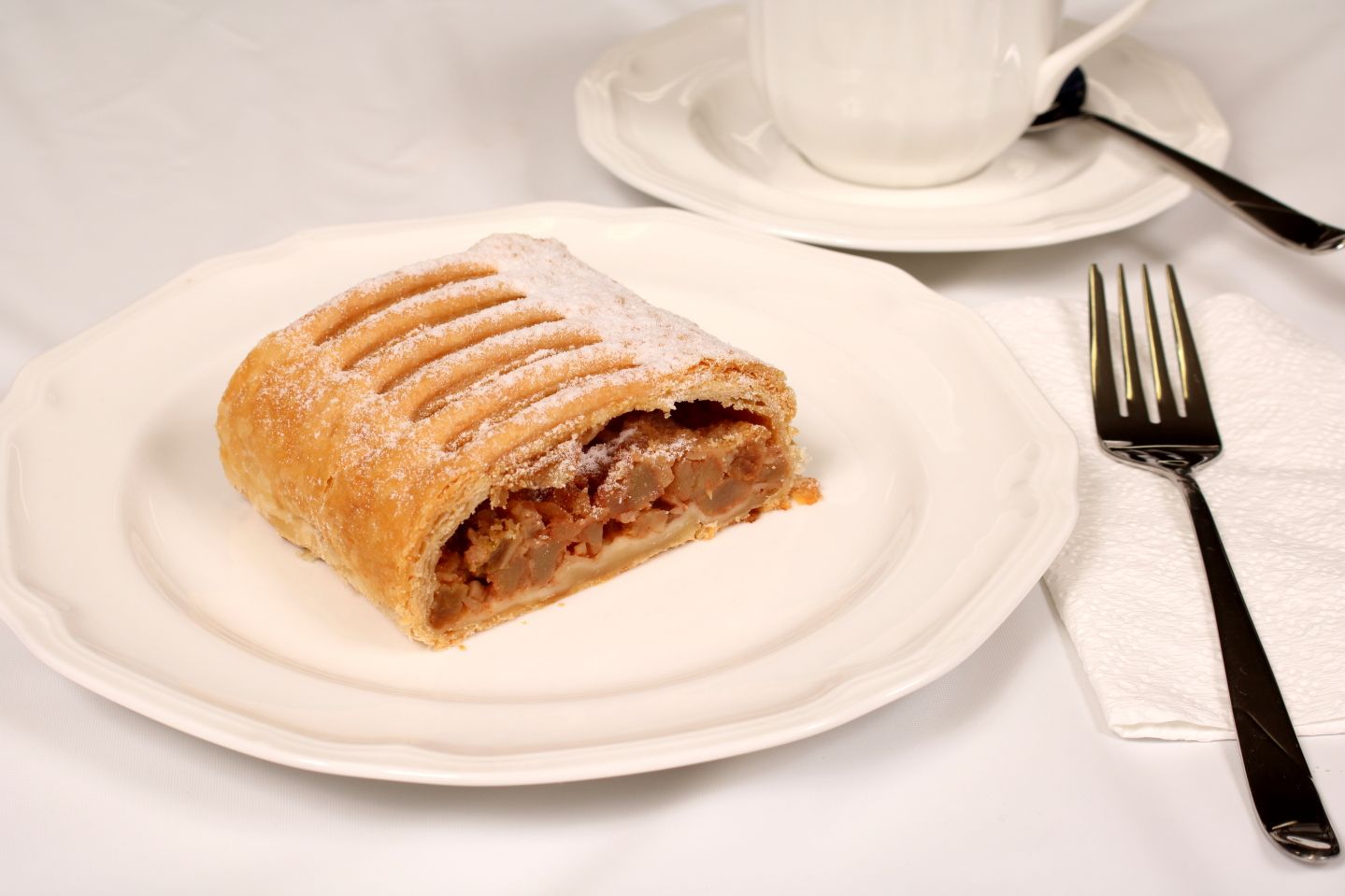 Apple strudel on a white plate with a fork, a cup of tea, and a napkin.