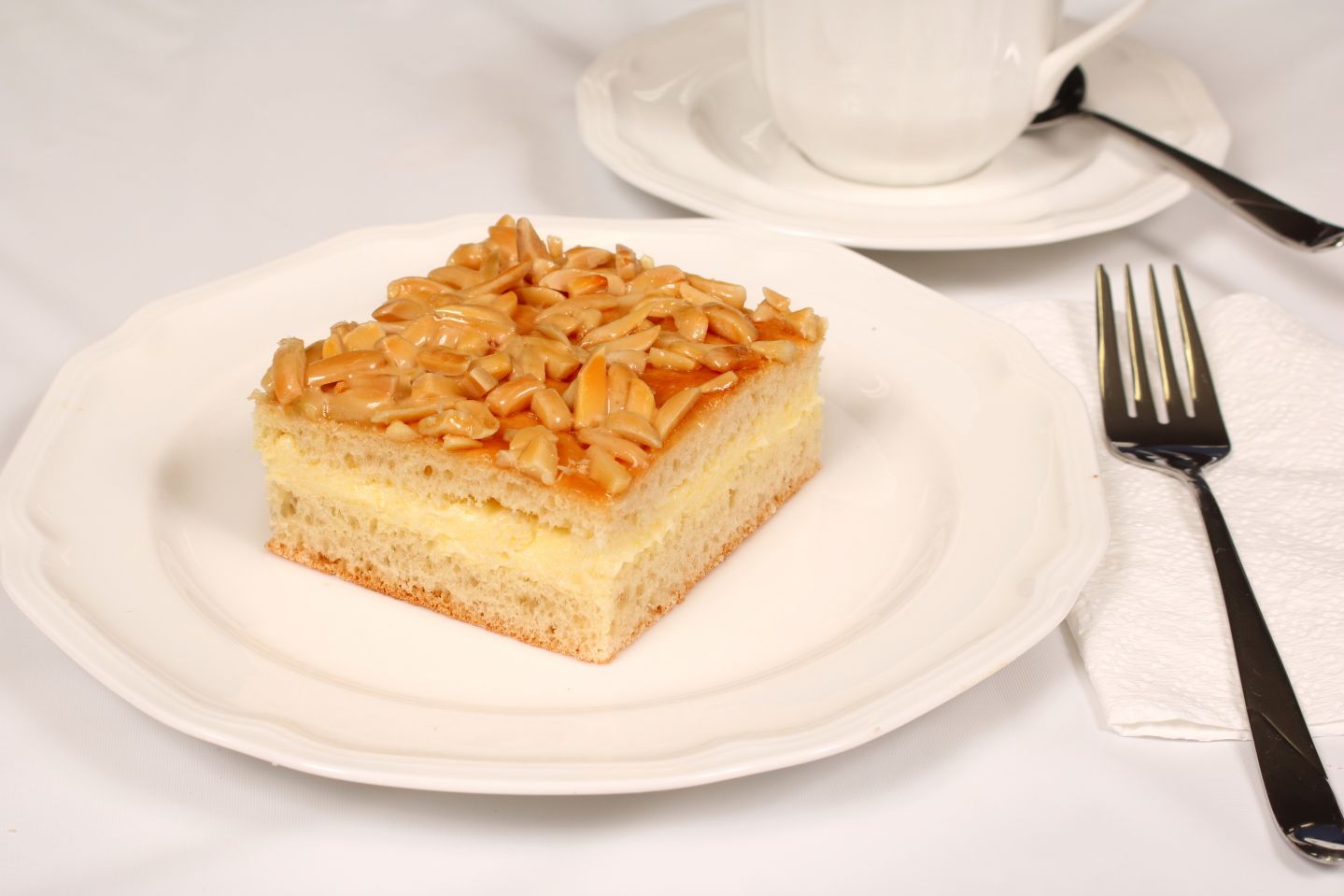Slice of cake with almond brittle topping on a white plate, fork, cup, and saucer.