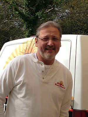 Man with beard and glasses smiles, standing next to a van. He wears a white shirt with a logo.