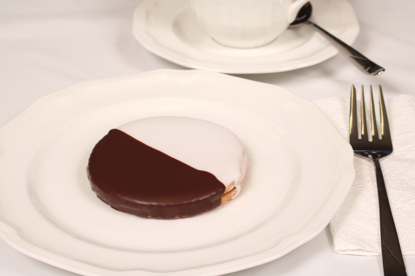 Black and white cookie on a white plate with a teacup and fork.