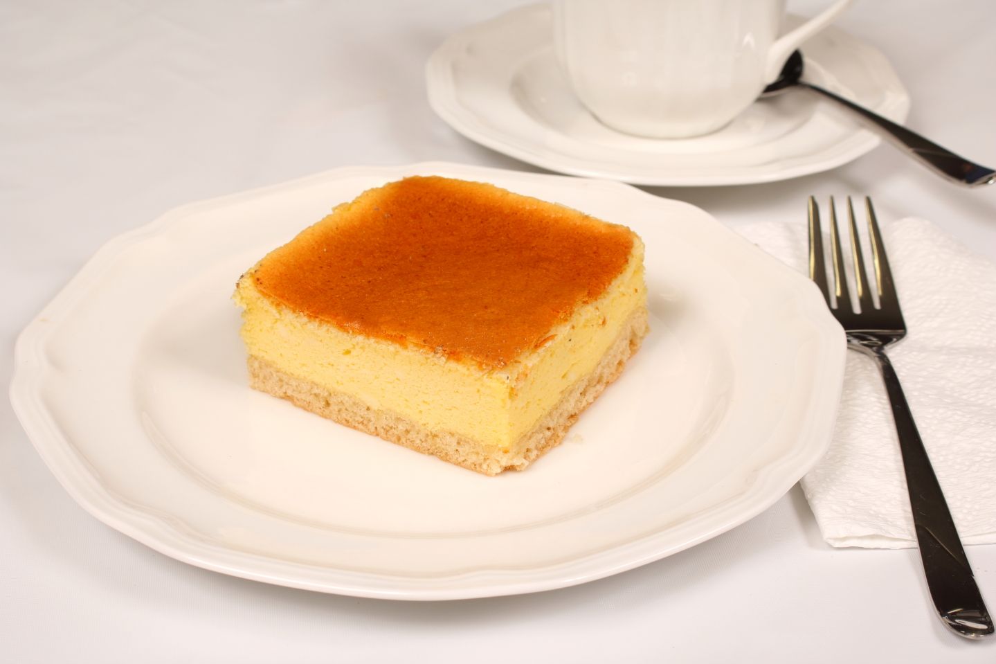 Cheesecake square on white plate, beside fork, with teacup in the background.