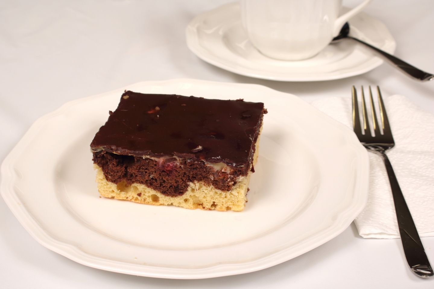 Slice of cake with chocolate frosting on white plate, beside cup, saucer, fork, and napkin.