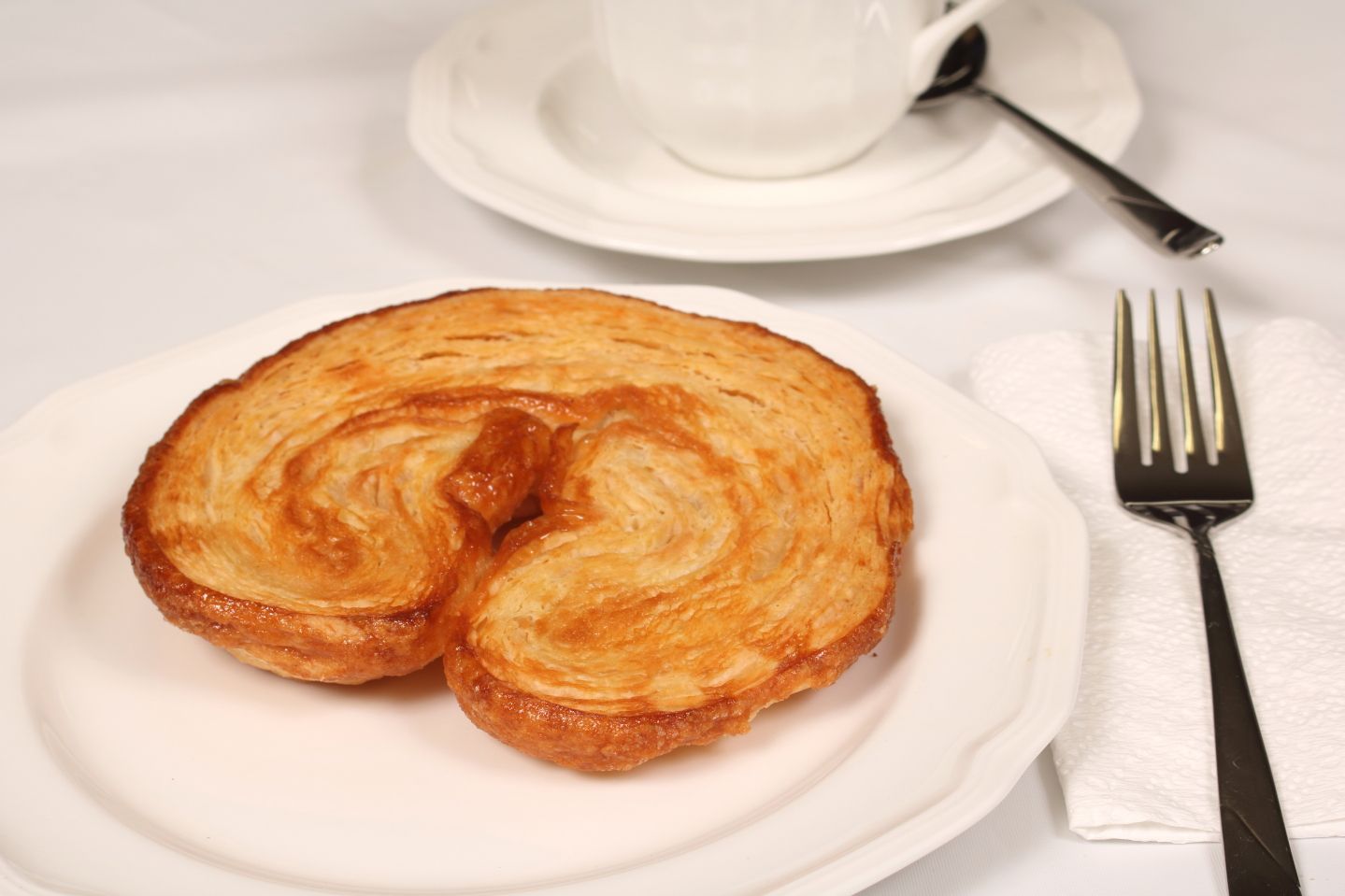 Palmier pastry on a white plate with a fork, a cup, saucer, and napkin.