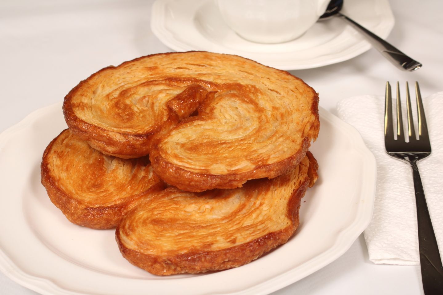 Three palmiers pastries on a white plate with a fork, a cup and saucer in background.