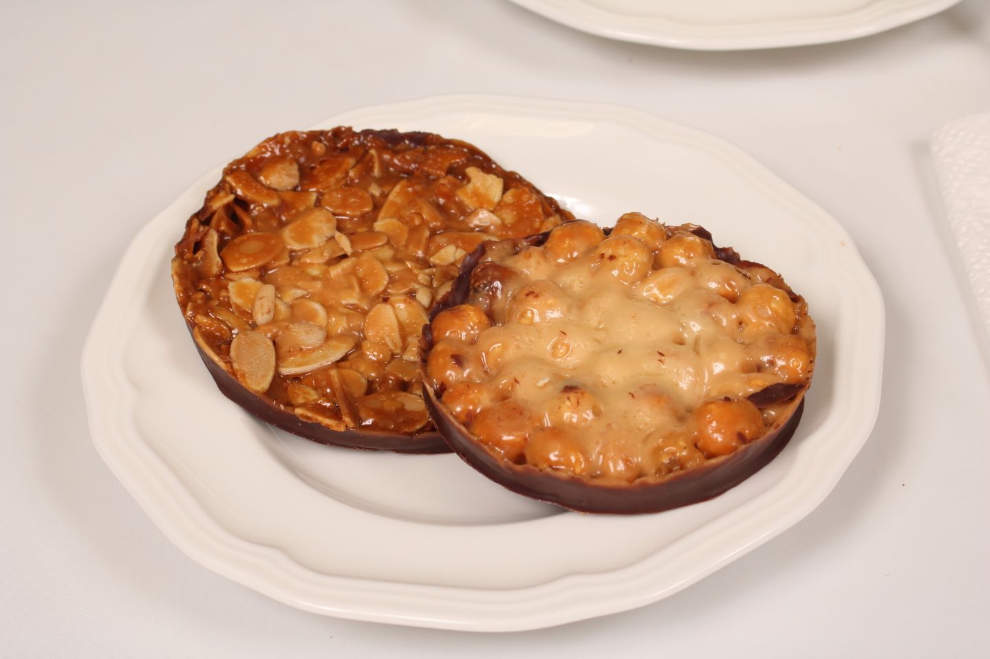 Two round, brown pastries on a white plate. One topped with sliced almonds, the other with caramelized nuts.
