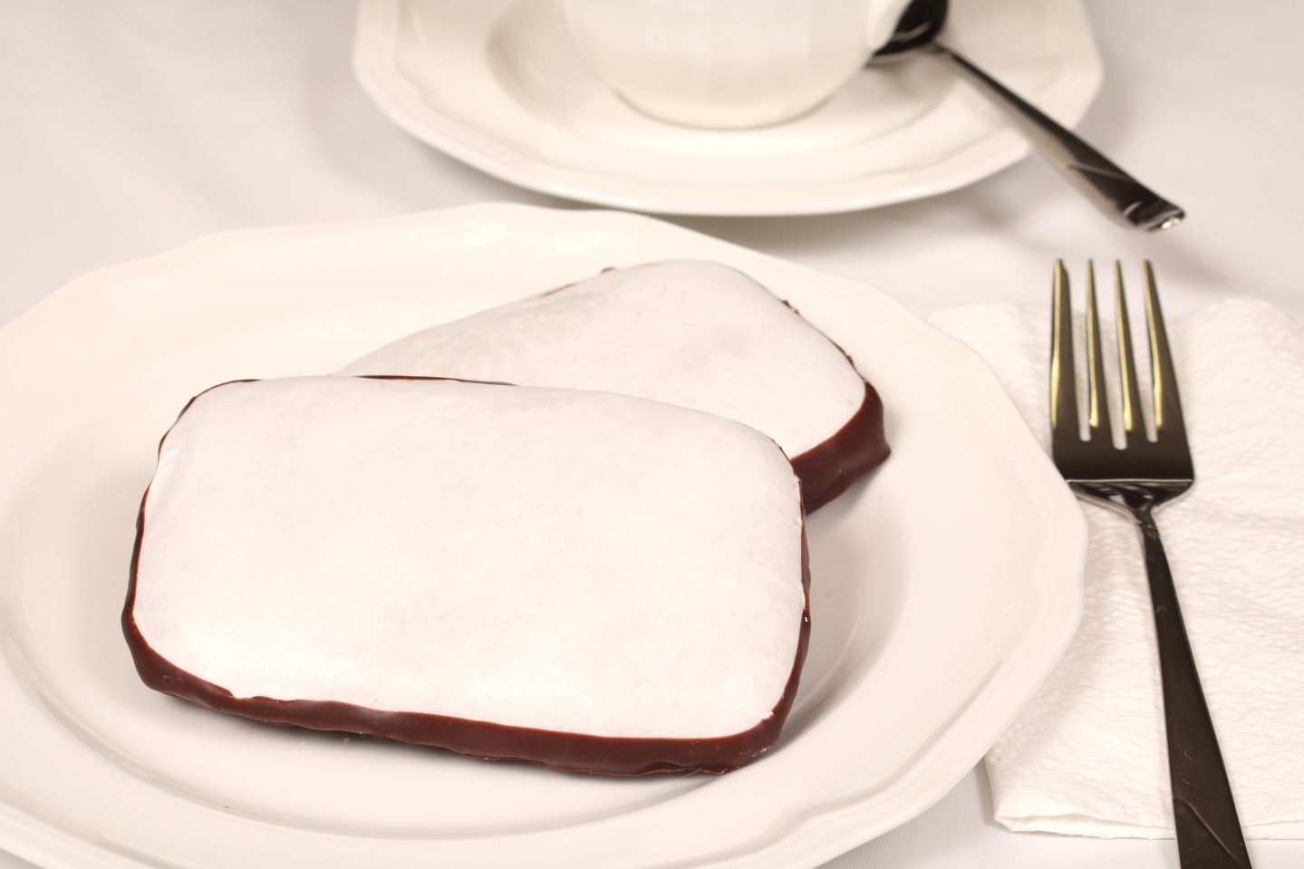 Two white-frosted, chocolate-covered cakes on a white plate, with a fork, napkin, and teacup in the background.