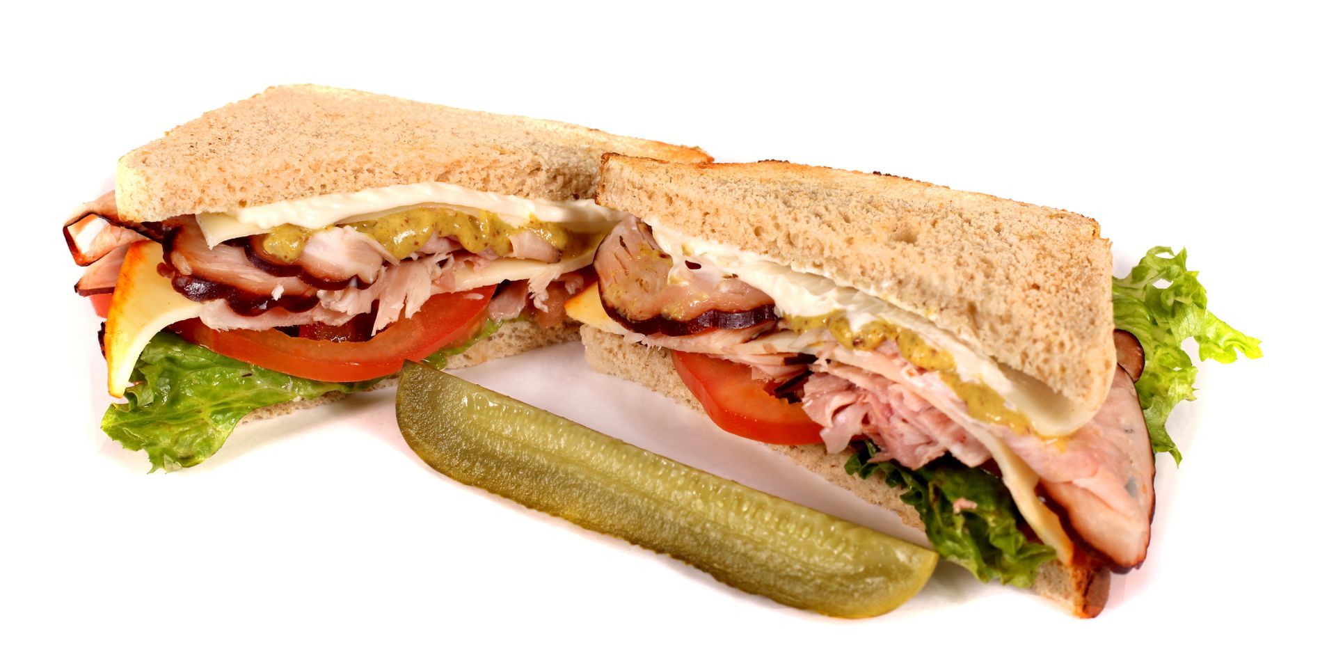 Sandwich cut in half, with ham, cheese, tomato, lettuce, and a pickle on a white surface.