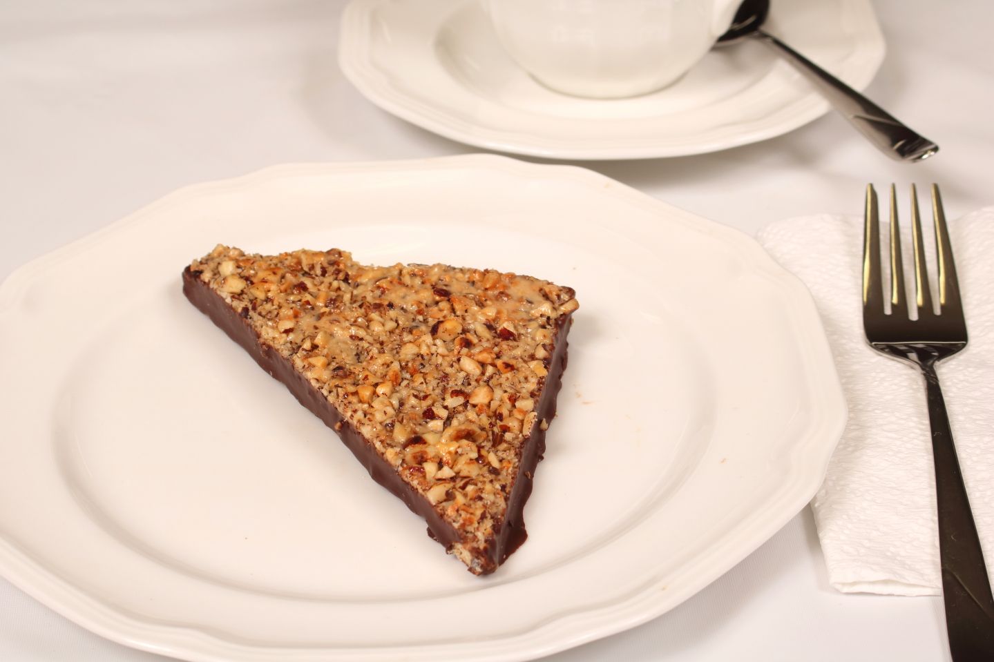 Slice of chocolate pecan pie on a white plate with fork, napkin, and teacup in a bright setting.