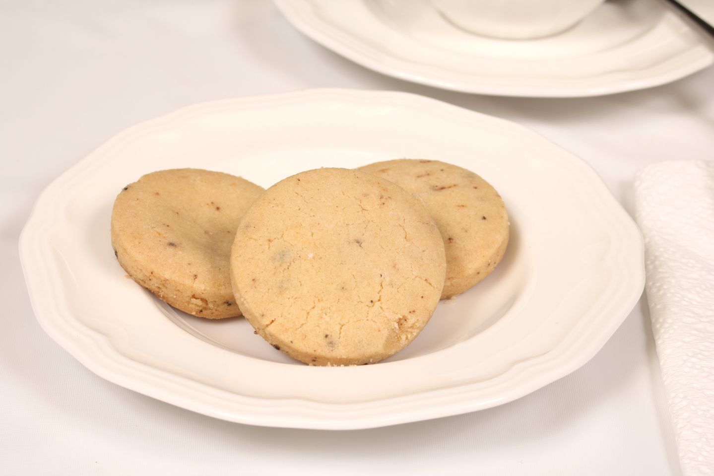 Three round cookies on a white plate, with a teacup and napkin in the background.