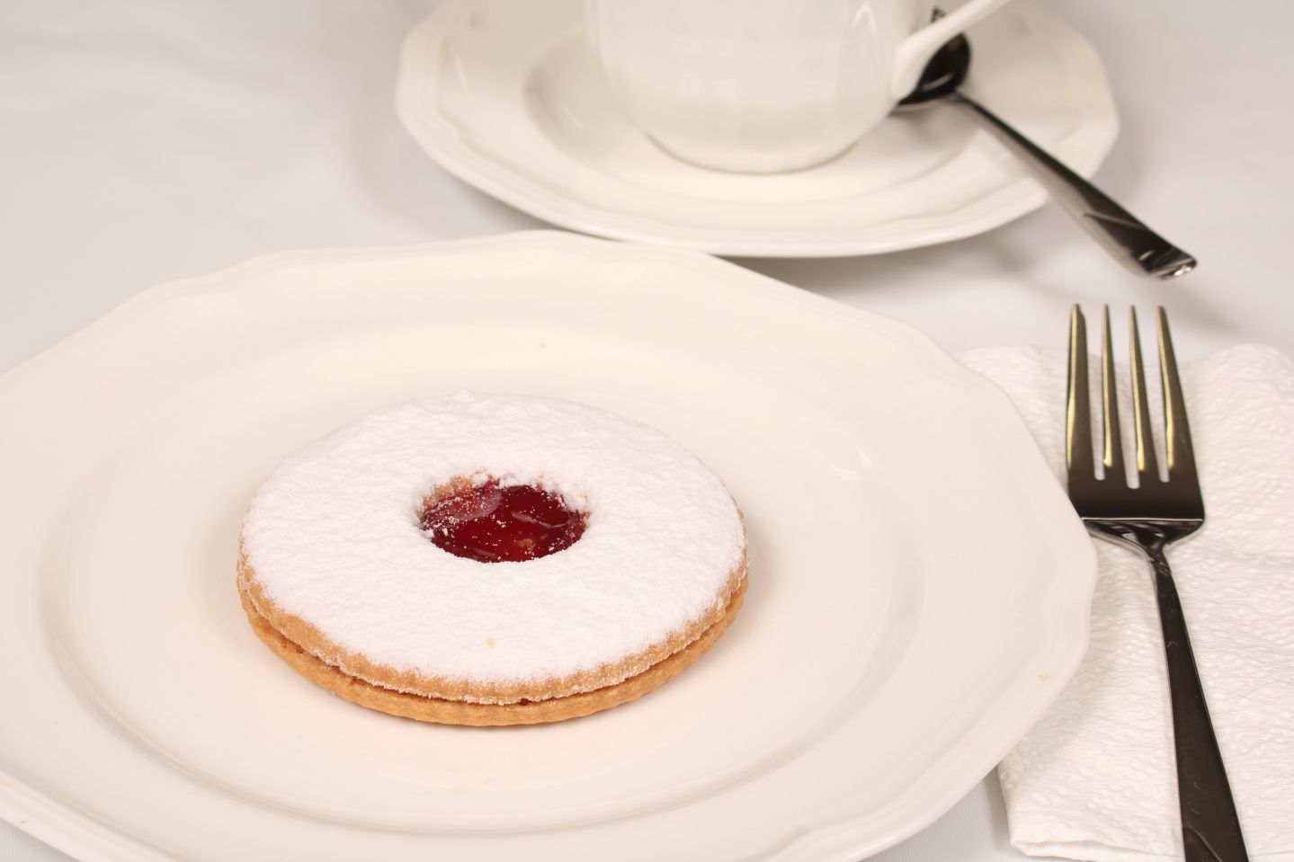 A shortbread cookie with jam center dusted with powdered sugar on a white plate, with a fork, a cup, and saucer.