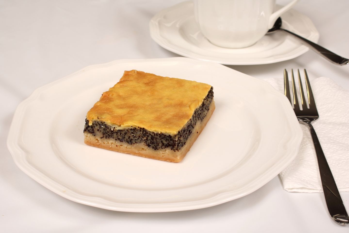 Square poppy seed cake on a white plate with a fork, napkin, and teacup in the background.