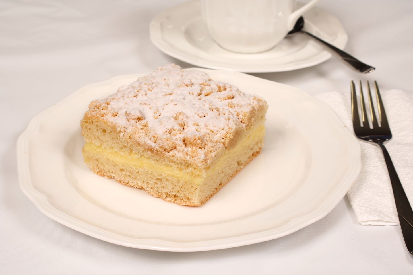 Cake slice on plate, dusted with powdered sugar, next to a fork, with a teacup in the background.