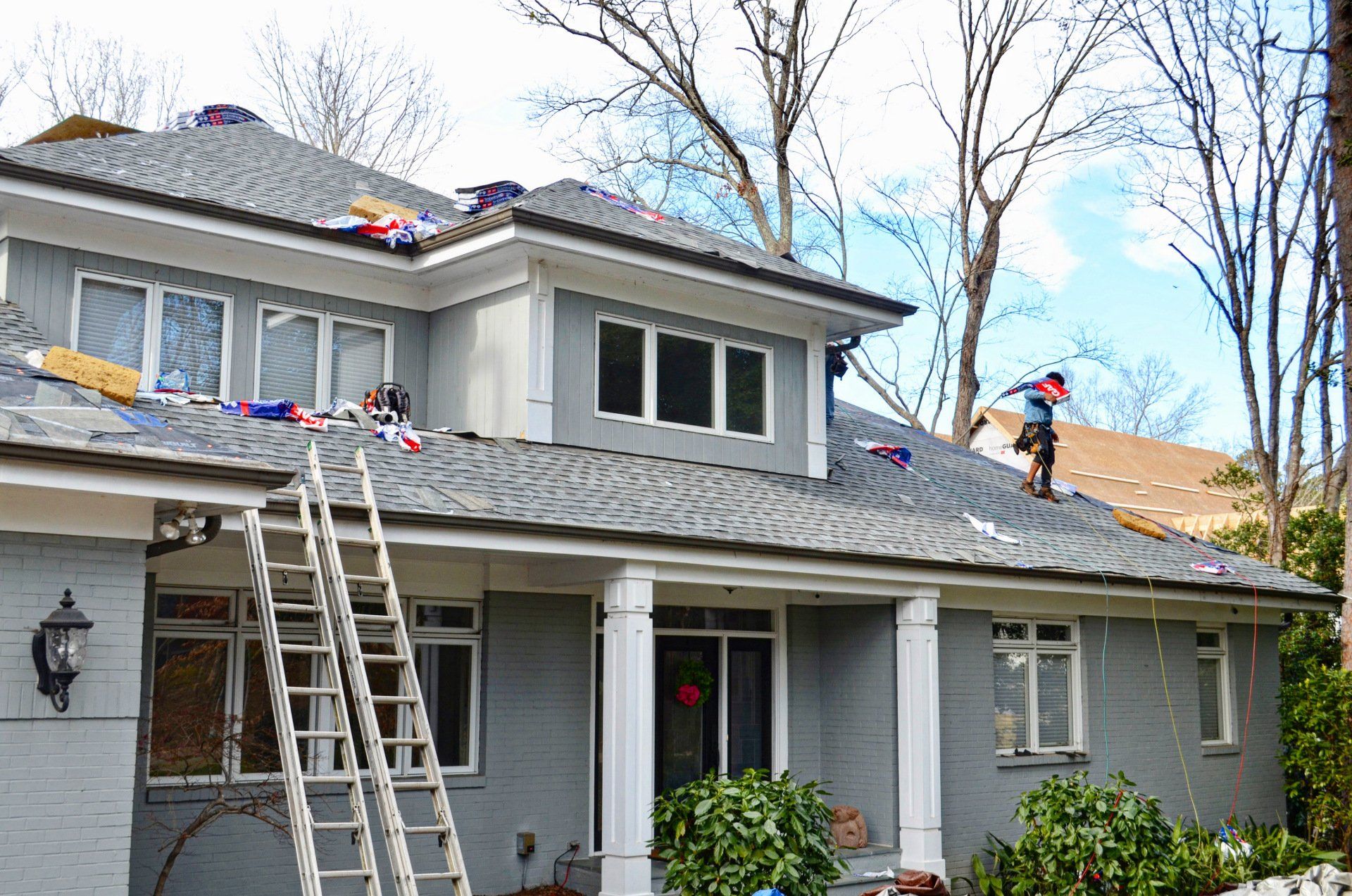 Roofers in Raleigh installing new shingles on a two-story gray home with ladders and tools on the roof and ground.