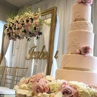A tiered pink wedding cake with flower decorations, next to a mirror with floral arrangements and the name 
