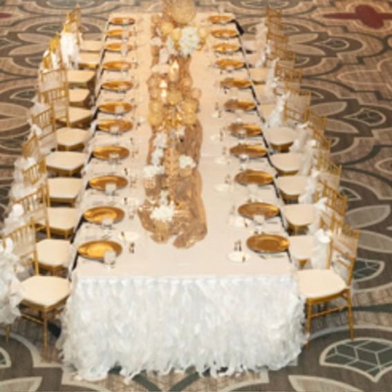 Long banquet table set for a formal event. Gold plates and chairs, white linens, and gold centerpieces.