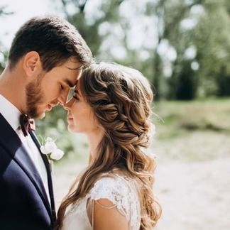 Bride and groom touch foreheads, side view. He wears a navy suit, she wears a white dress; outdoor setting.