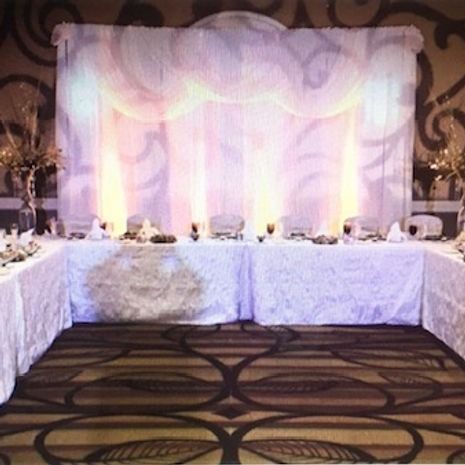 Head table set up at a wedding reception, with white draping and lighting; a floral patterned carpet.