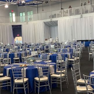Blue-tabled banquet hall, white drapes, silver chairs. Tables set for dining. Large windows and balcony.
