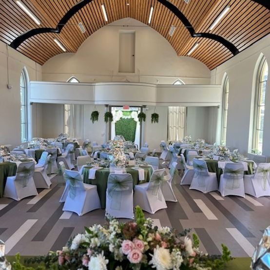 Wedding reception setup with round tables, white chairs, green linens, and floral centerpieces in a light-filled hall.