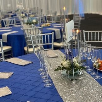 Tables set for an event: blue tablecloths, silver chairs, centerpieces, and glassware.