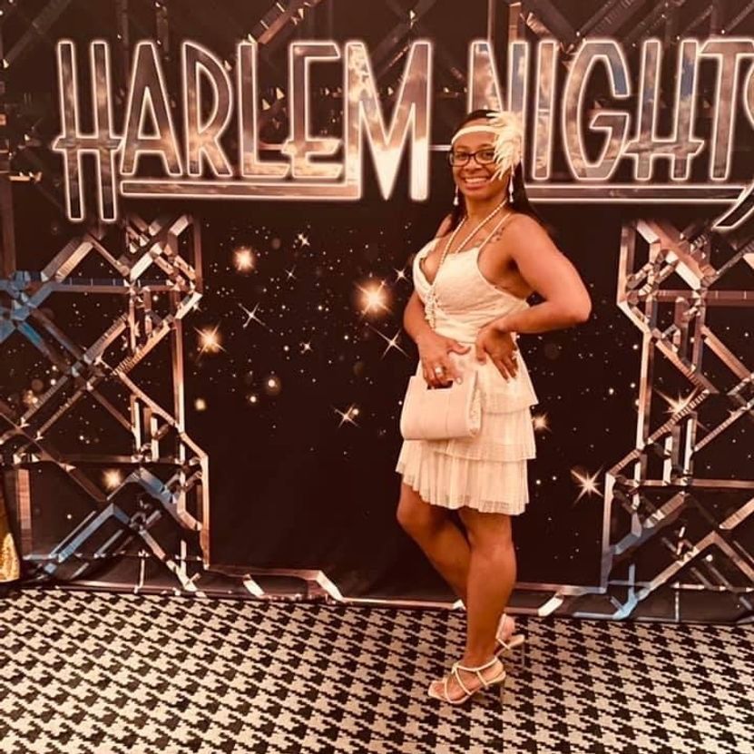 Woman in flapper dress, headband, and purse poses in front of a 