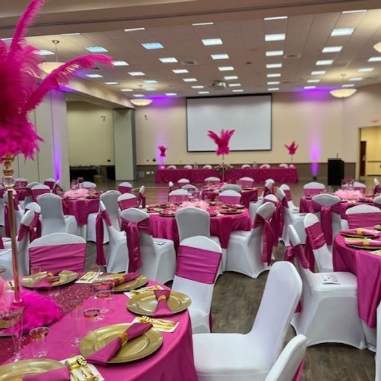 Pink and gold event decor: tables with pink tablecloths, white chairs, and feather centerpieces in a large room.