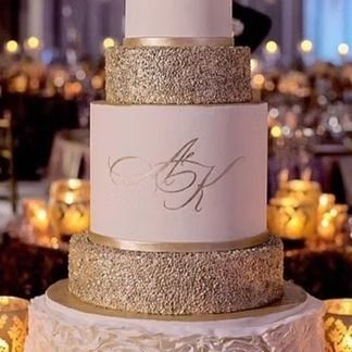 Four-tier wedding cake with gold glitter and initials, set on a table with candles.