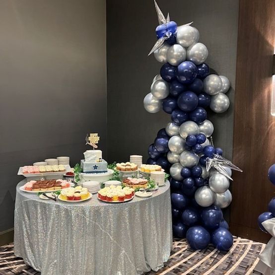 A dessert table with cake and treats, decorated in blue and silver with a balloon arch.