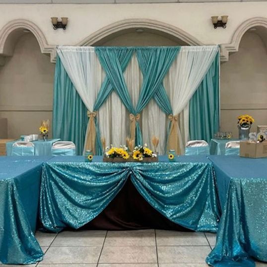 A turquoise sequined head table with a teal and white draped backdrop decorated with sunflowers.
