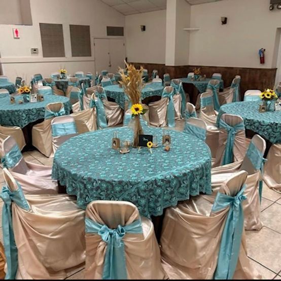 Round tables set for a party. Teal tablecloths, cream chairs with teal bows. Sunflower centerpieces.