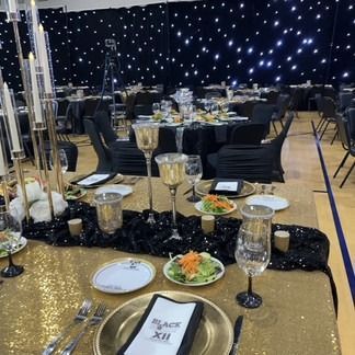 Elegant banquet hall with gold and black tables set for dinner, with starry backdrop.