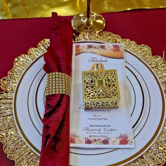 Formal table setting: gold charger, white plate, maroon napkin, gold napkin ring, favor box, menu.