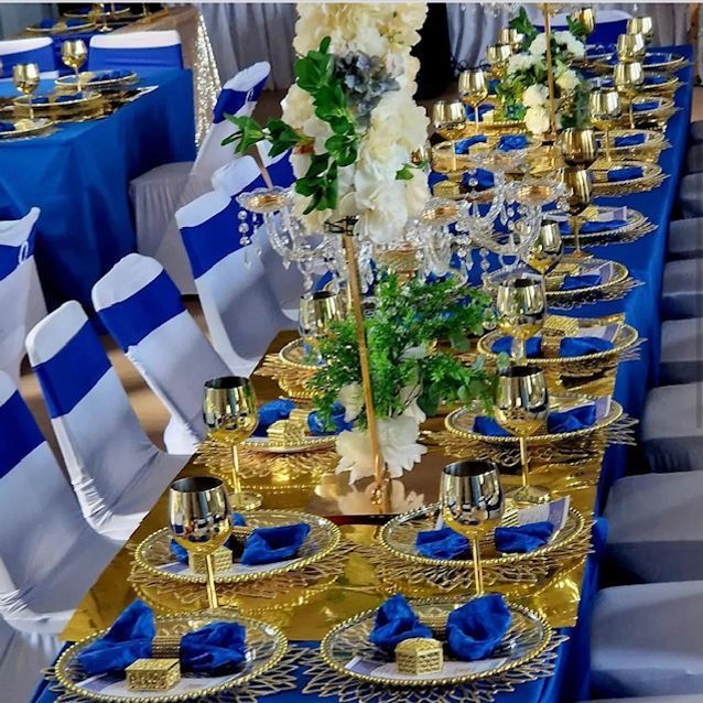 Long banquet table with blue and gold place settings, white and blue chair covers, floral centerpiece.
