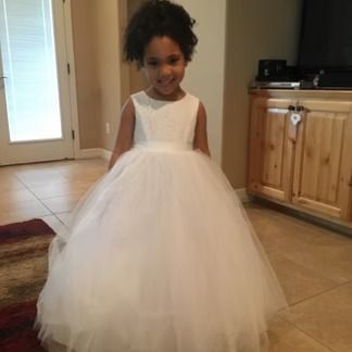 Young girl with curly hair smiles, wearing a white dress in a home setting.