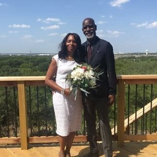 Couple stands on a wooden deck overlooking trees, woman in white dress holds flowers, man in suit.