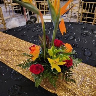 Floral centerpiece: Bird of paradise flowers, red roses, yellow lilies on gold runner, black tablecloth.