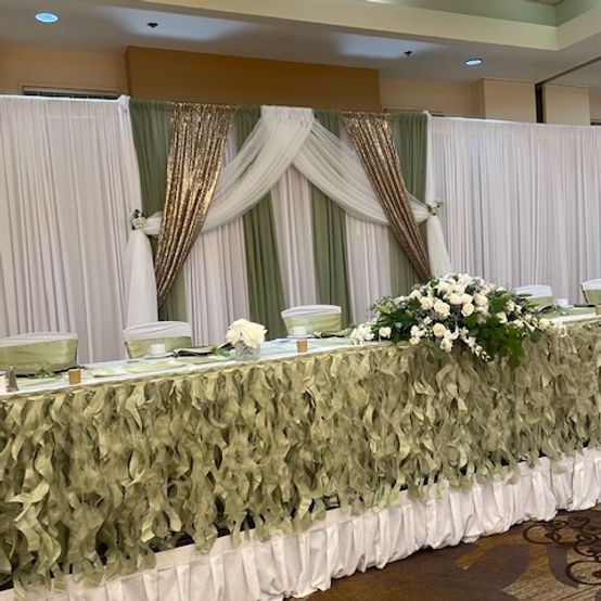 Wedding reception head table with white and gold draped backdrop and sage green floral garland.