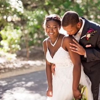 Smiling bride in white dress with groom kissing her shoulder; outdoor setting.