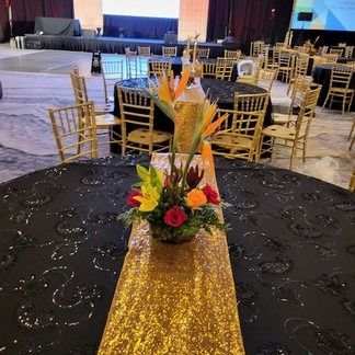 Round table with gold and black tablecloth. Floral centerpiece with Bird of Paradise, setting at an event.
