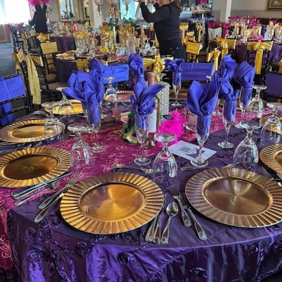Round table set for a party with gold chargers, purple linens, blue napkins, and a woman arranging decorations.