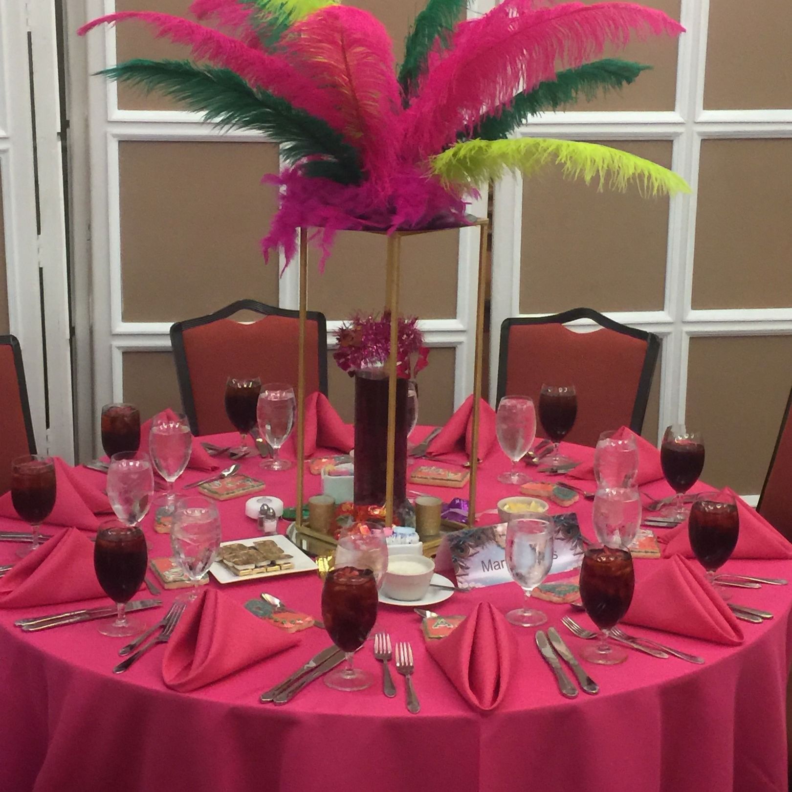 Table setting for an event with hot pink tablecloth, napkins, and red glasses. Gold centerpiece and feathers.