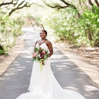 Bride in a white gown smiles, holding bouquet, standing on a road lined with trees.
