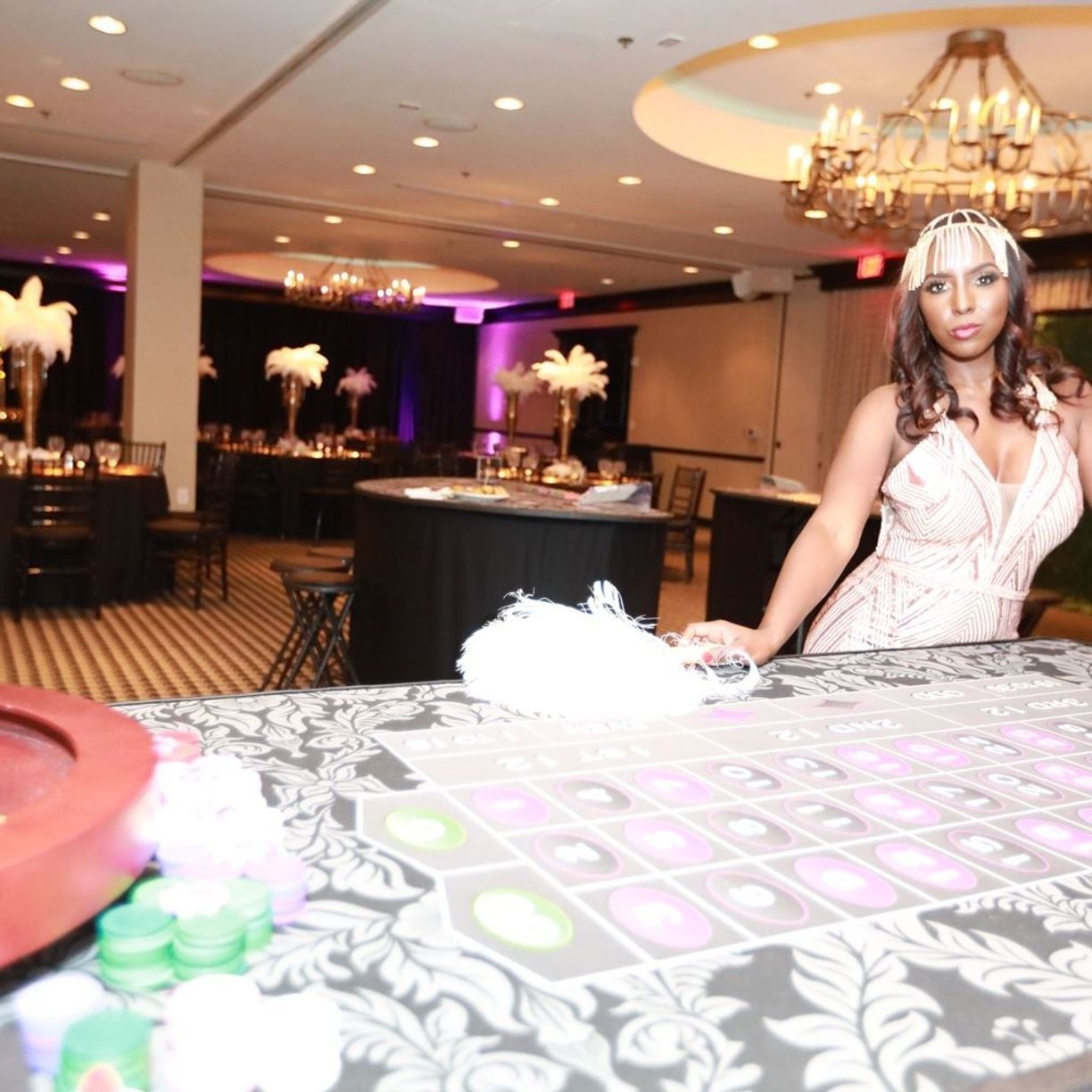 Woman in a flapper dress at a roulette table in a casino-themed event with decorations and chandeliers.
