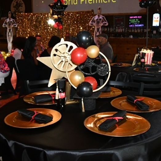 A table setting with a movie theme: gold plates, black napkins, film reel centerpiece, and balloons.