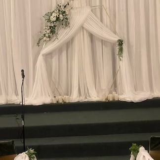 Wedding ceremony backdrop with sheer white fabric, geometric arch, and floral decorations.