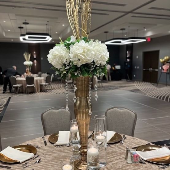 Elegant table centerpiece: gold vase with white flowers, tall gold branches.  Setting: ballroom, table with place settings.