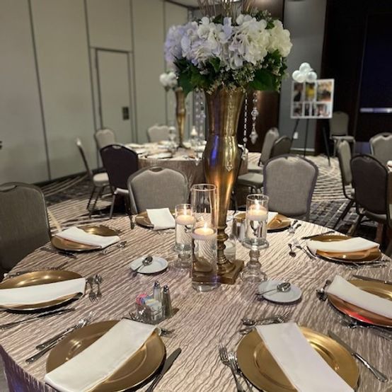 Round table set for a formal event with gold accents, tall floral centerpiece, and candles.